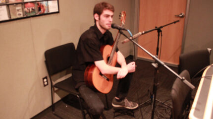 A teen holding an acoustic guitar sitting behind a microphone in a radio studio