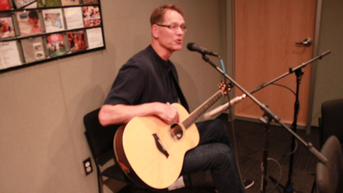 An adult playing acoustic guitar and singing into a microphone in a radio studio