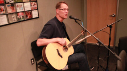 An adult playing acoustic guitar and singing into a microphone in a radio studio