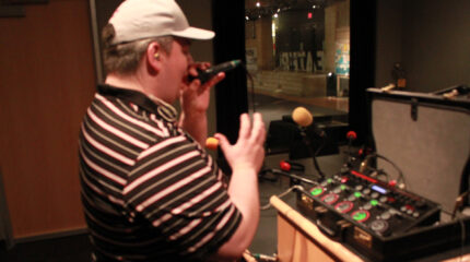An adult singing into a microphone in front of a suitcase with an audio mixing device inside in a radio studio