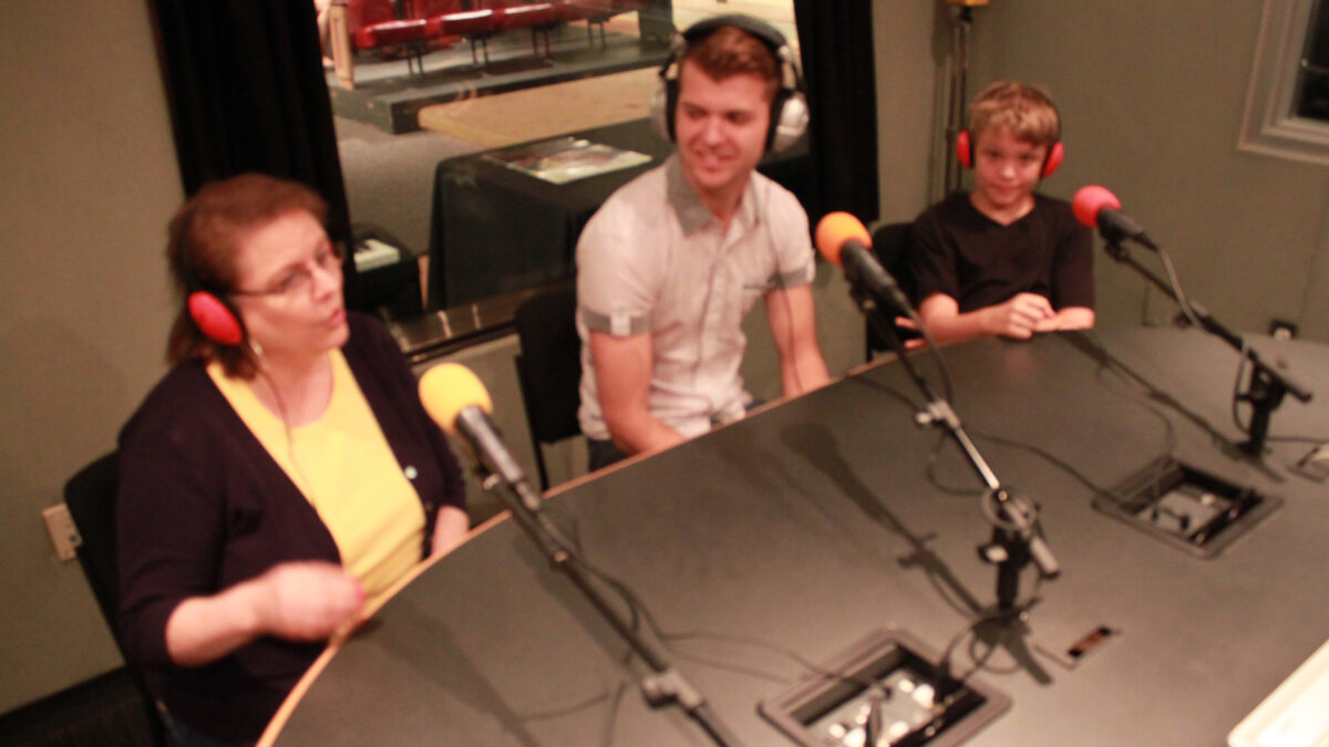 Two adults and a youth wearing headphones and speaking into microphones in a radio studio