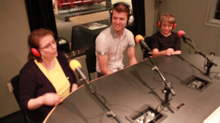 Two adults and a youth wearing headphones and speaking into microphones in a radio studio