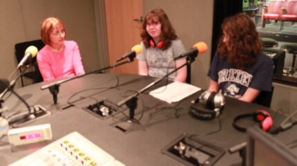 An adult and two teens speaking into headphones in a radio studio