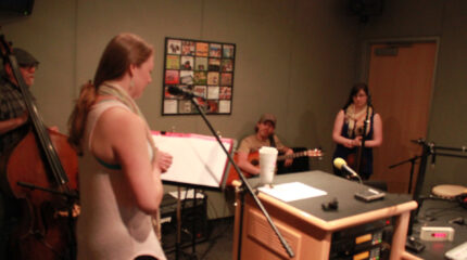 An adult holding an upright bass, an adult standing behind sheet music on a music stand and a microphone, an adult sitting while holding an acoustic guitar, and an adult standing behind a microphone in a radio studio