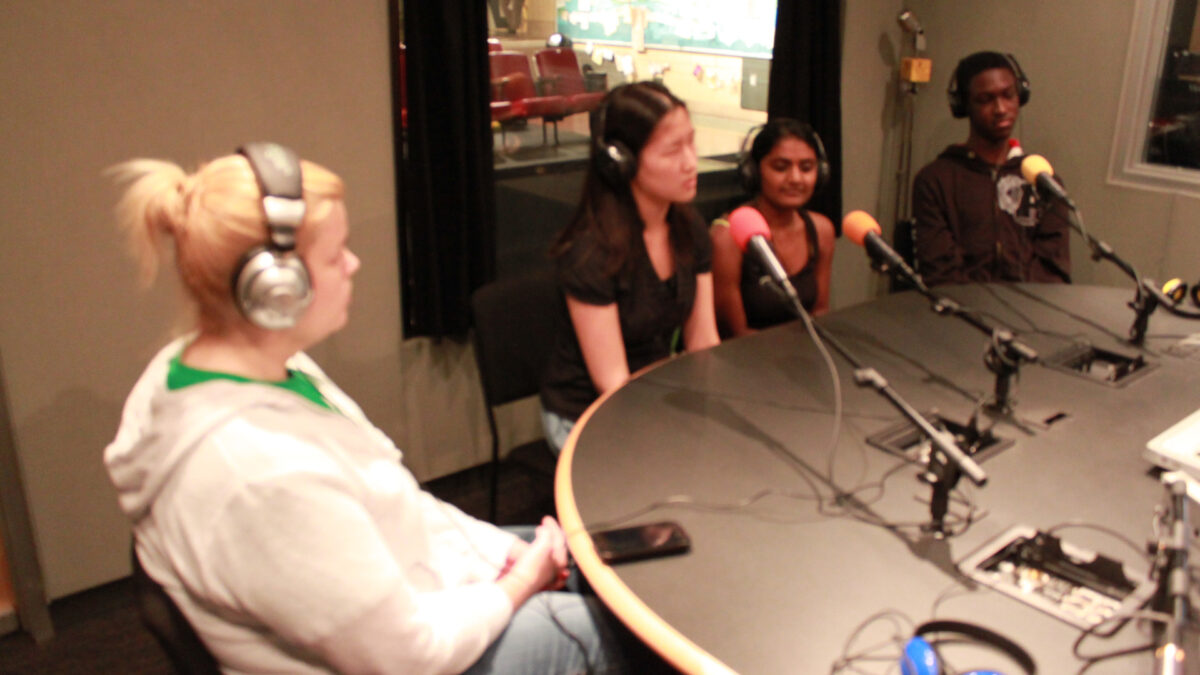 Three teens and an adult wearing headphones speaking into microphones in a radio studio