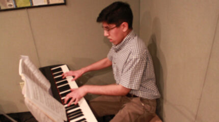 A teen playing a piano keyboard looking at sheet music in a radio studio.