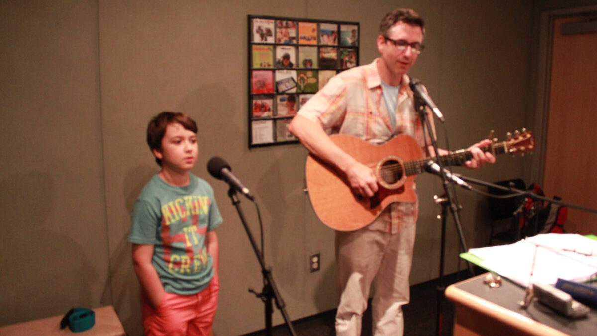 A youth singing into a microphone and an adult playing an acoustic guitar and singing into a microphone in a radio studio