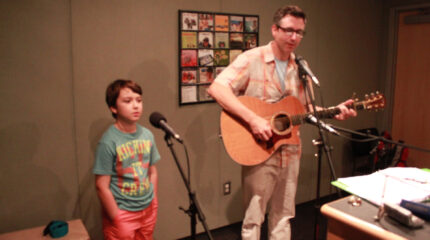 A youth singing into a microphone and an adult playing an acoustic guitar and singing into a microphone in a radio studio