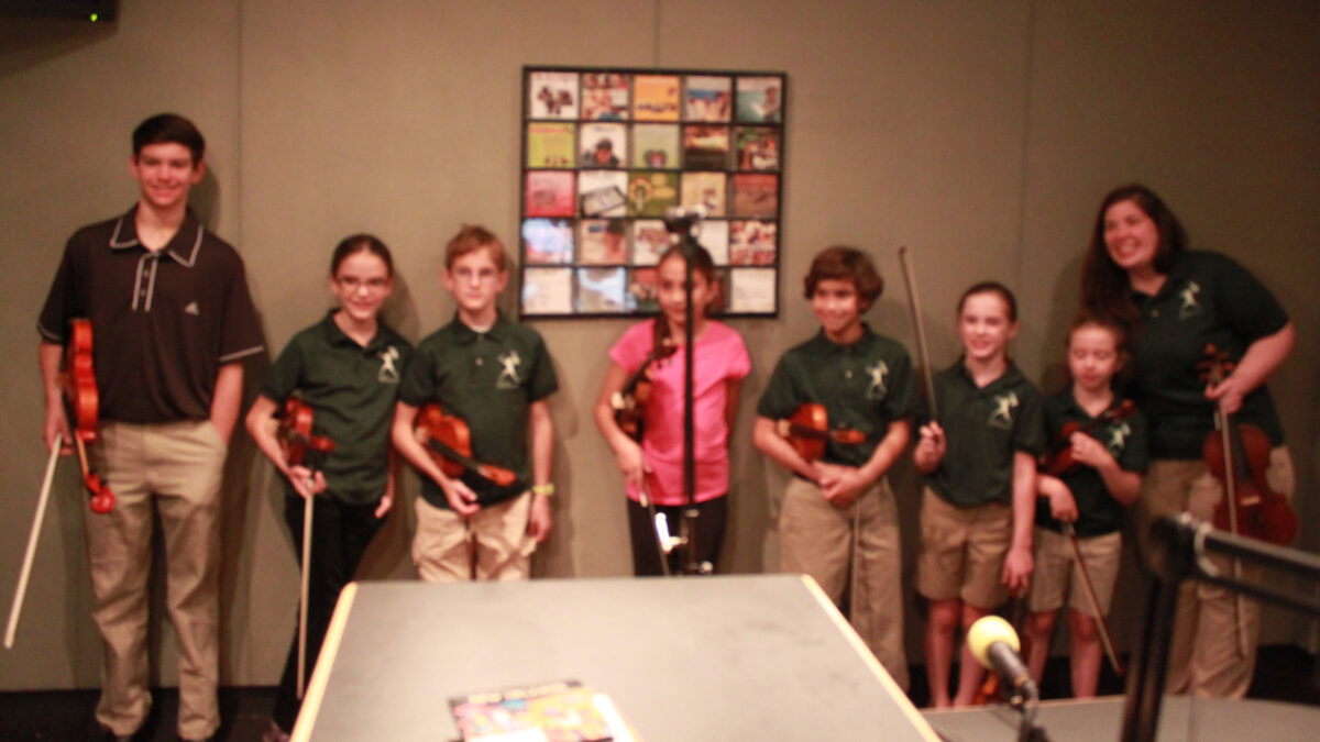 Six youth and two teens holding violins and smiling in a radio studio