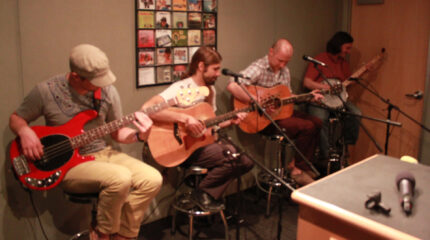 An adult playing electric bass, two adults playing acoustic guitars, and an adult playing banjo in a radio studio