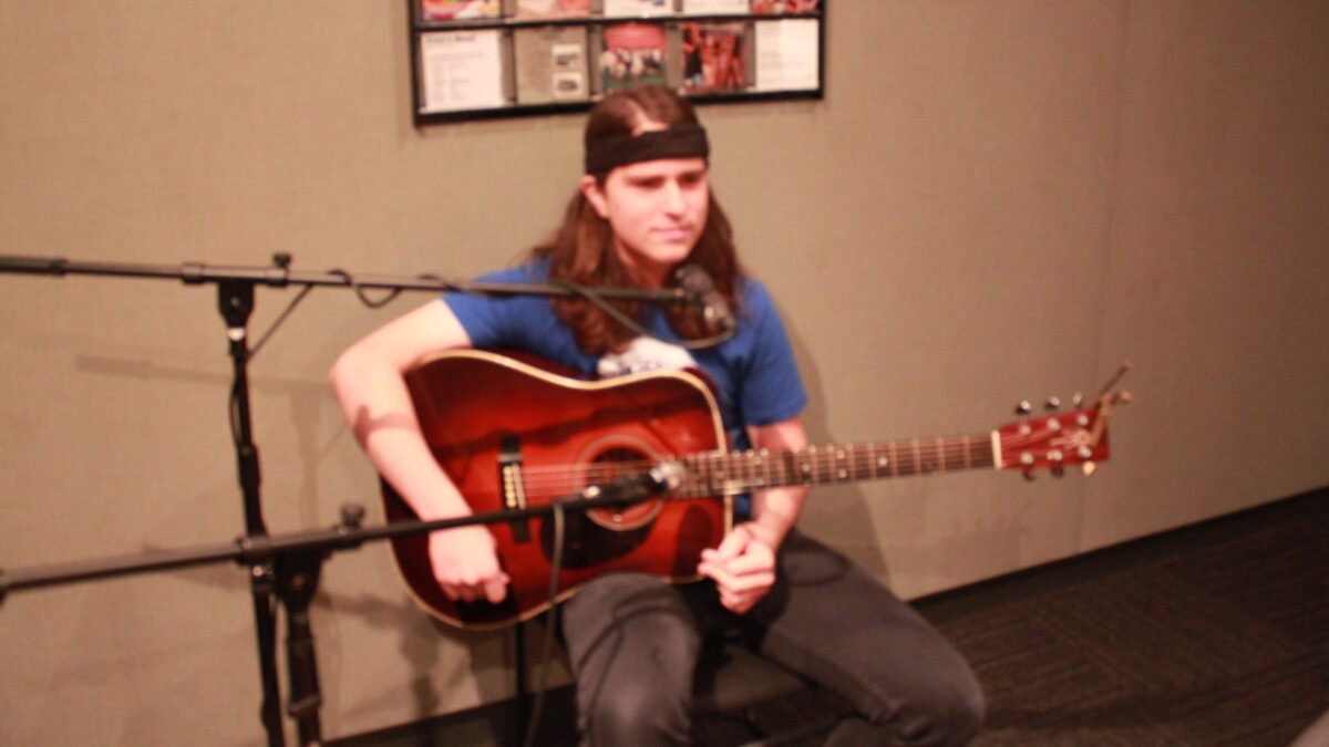 A teen holding an acoustic guitar and sitting behind a microphone in a radio studio