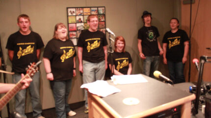 Six adults singing in a choir in a radio studio