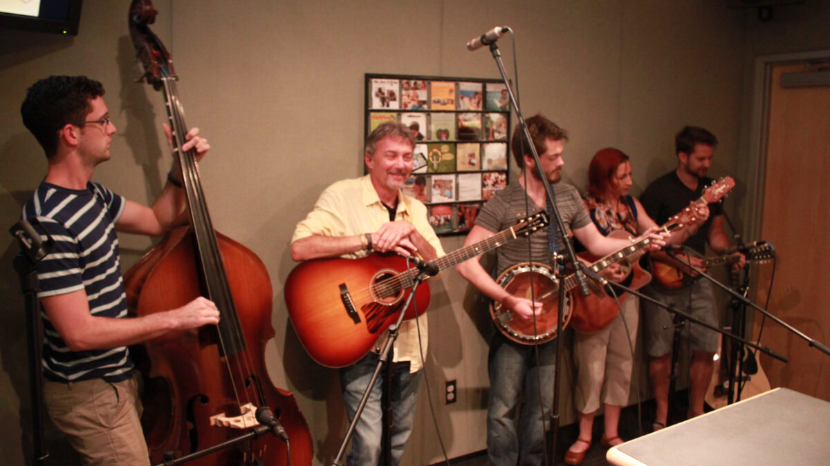 An adult playing an upright bass, an adult holding an acoustic guitar and smiling, an adult playing a banjo, an adult playing an acoustic guitar, and an adult playing a mandolin in a radio studio