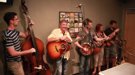 An adult playing an upright bass, an adult holding an acoustic guitar and smiling, an adult playing a banjo, an adult playing an acoustic guitar, and an adult playing a mandolin in a radio studio