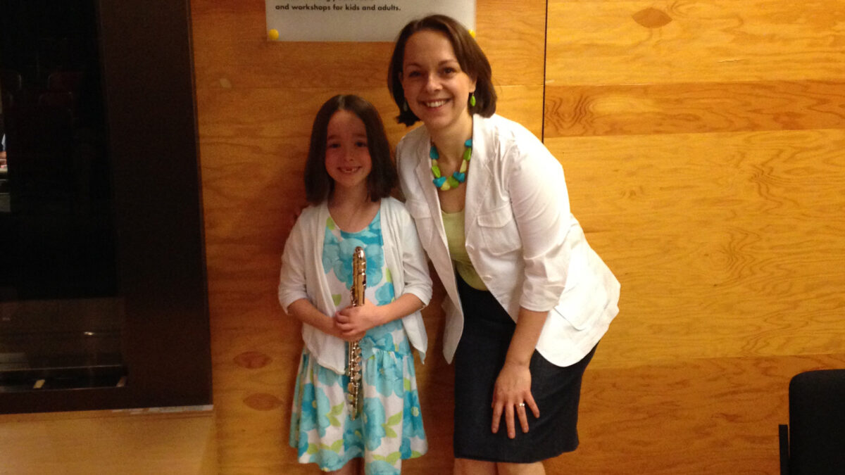 An adult smiling at the camera and a youth holding an alto flute and smiling at the camera