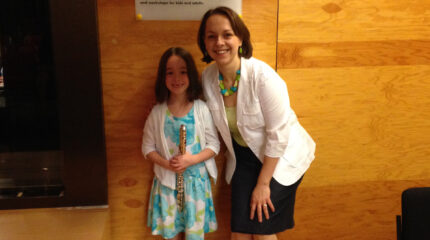 An adult smiling at the camera and a youth holding an alto flute and smiling at the camera