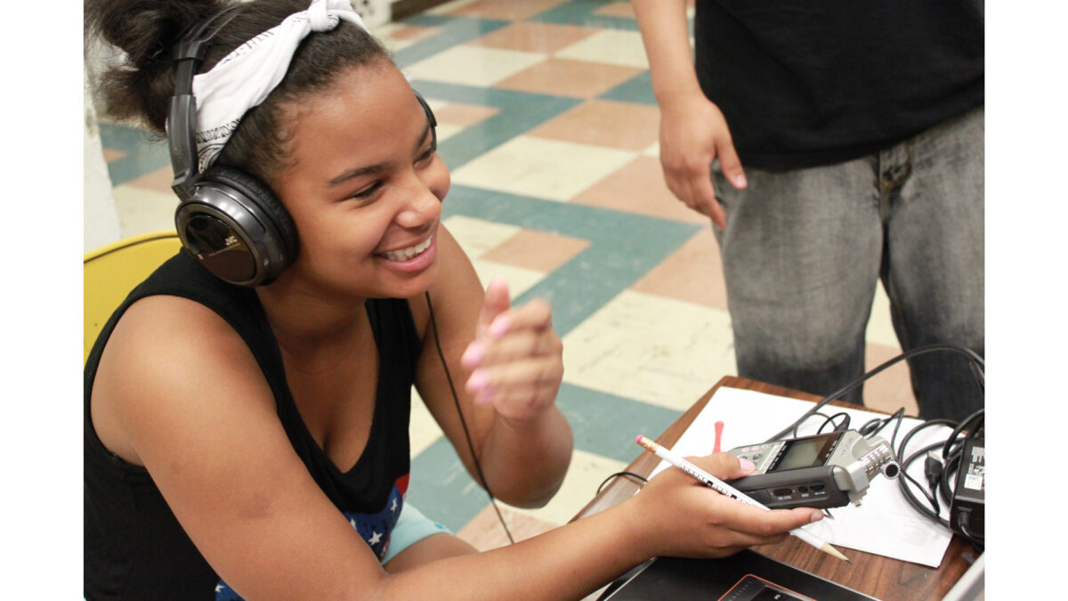 A youth wearing headphones smiling while holding a pencil and a portable recorder and sitting at a classroom desk