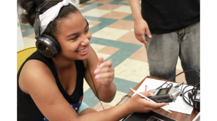 A youth wearing headphones smiling while holding a pencil and a portable recorder and sitting at a classroom desk