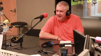 An adult wearing headphones speaking into a microphone in a radio studio