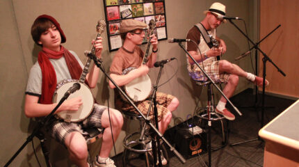 Two teens and an adult playing banjos while sitting behind microphones in a radio studio