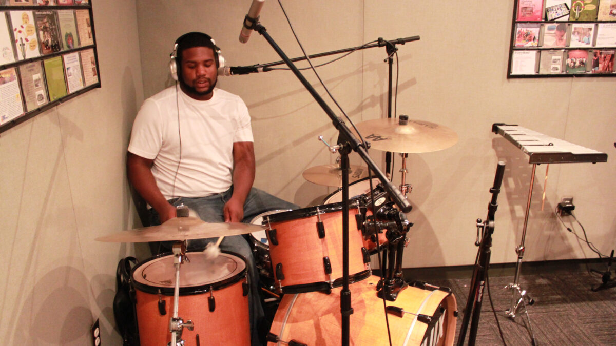 An adult wearing headphones playing a drum set in a radio studio