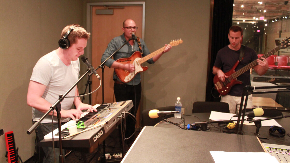 An adult wearing headphones playing a piano keyboard, an adult playing an electric guitar and singing into a microphone, and an adult playing an electric bass in a radio studio
