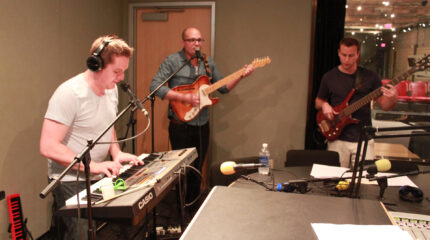 An adult wearing headphones playing a piano keyboard, an adult playing an electric guitar and singing into a microphone, and an adult playing an electric bass in a radio studio