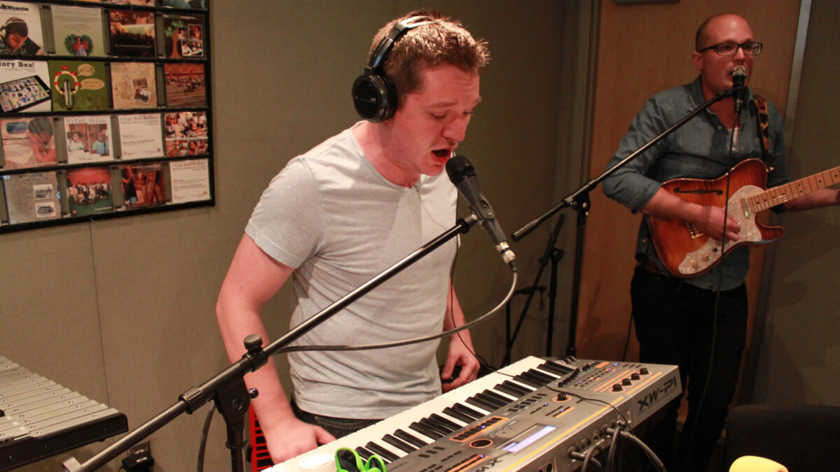 An adult standing behind a piano keyboard and singing into a microphone and an adult playing an electric guitar and singing into a microphone in a radio studio