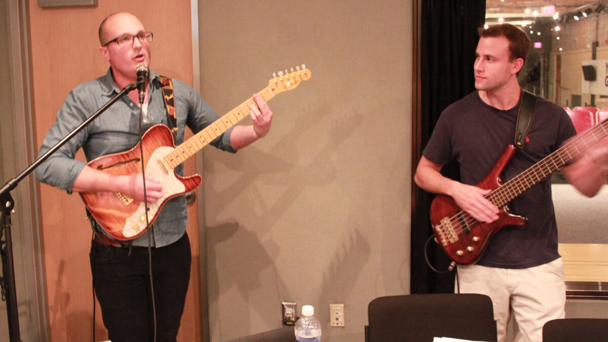 An adult playing electric guitar and singing into a microphone and an adult playing electric bass in a radio studio