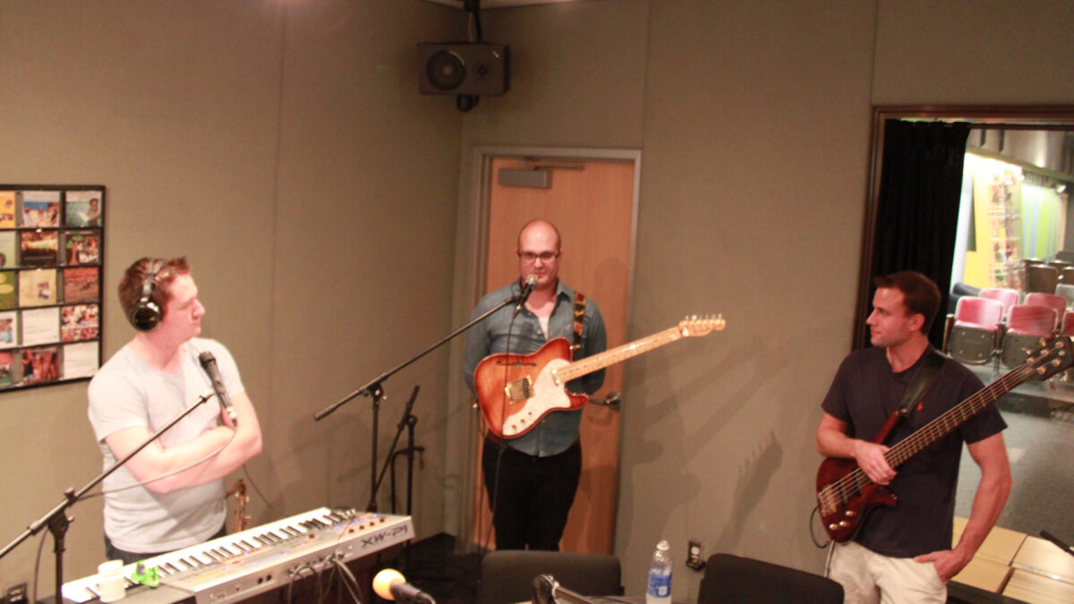 An adult wearing headphones standing behind a piano keyboard and a microphone, an adult holding an electric guitar standing behind a microphone, and an adult holding an electric bass in a radio studio