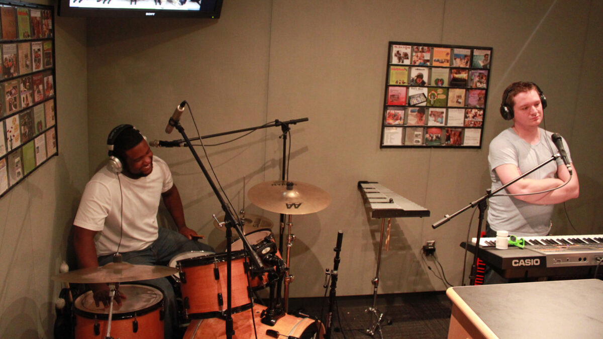 An adult wearing headphones sitting at a drum set and an adult wearing headphones standing behind a piano keyboard and a microphone next to a glockenspiel in a radio studio