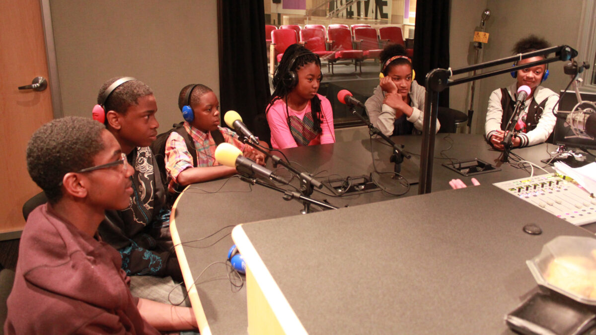 Six youth, five wearing headphones, sitting behind microphones in a radio studio