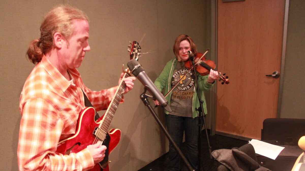 An adult playing an electric guitar and standing behind a microphone and an adult playing a violin and standing behind a microphone in a radio studio