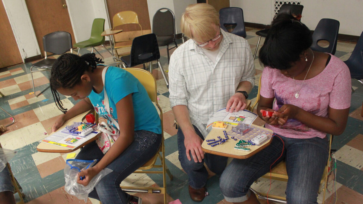 Two youth and an adult interacting with small electrical contraptions on classroom desks