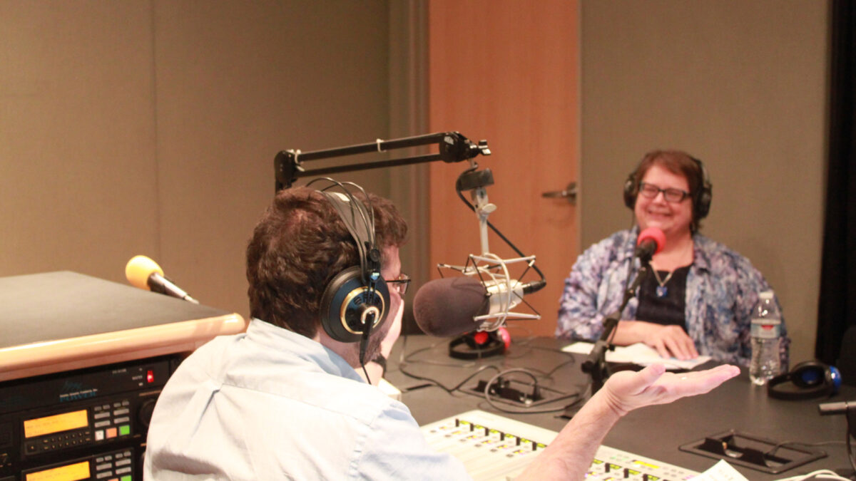An adult wearing headphones smiling while sitting behind a microphone being interviewed by an adult wearing headphones speaking into a microphone and sitting behind a mixer in a radio studio