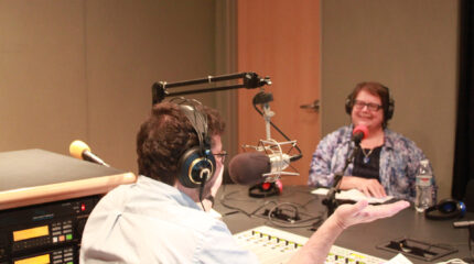 An adult wearing headphones smiling while sitting behind a microphone being interviewed by an adult wearing headphones speaking into a microphone and sitting behind a mixer in a radio studio