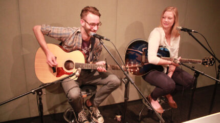 An adult playing acoustic guitar and singing into a microphone and an adult holding an acoustic guitar and sitting behind a microphone in a radio studio