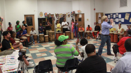 A large group of youth sitting behind African drums looking at an adult gesturing with their arms as an audience of adults watch in a classroom