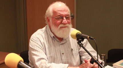 An adult smiling while sitting behind a microphone in a radio studio