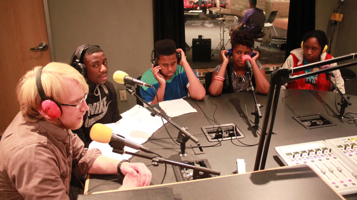Five teens wearing headphones speaking into microphones in a radio studio