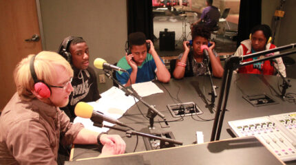 Five teens wearing headphones speaking into microphones in a radio studio