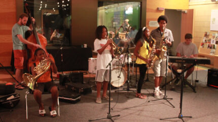 An adult playing upright bass, a youth playing french horn, a youth playing trumpet, two teens playing alto saxophone, and a teen playing a piano keyboard