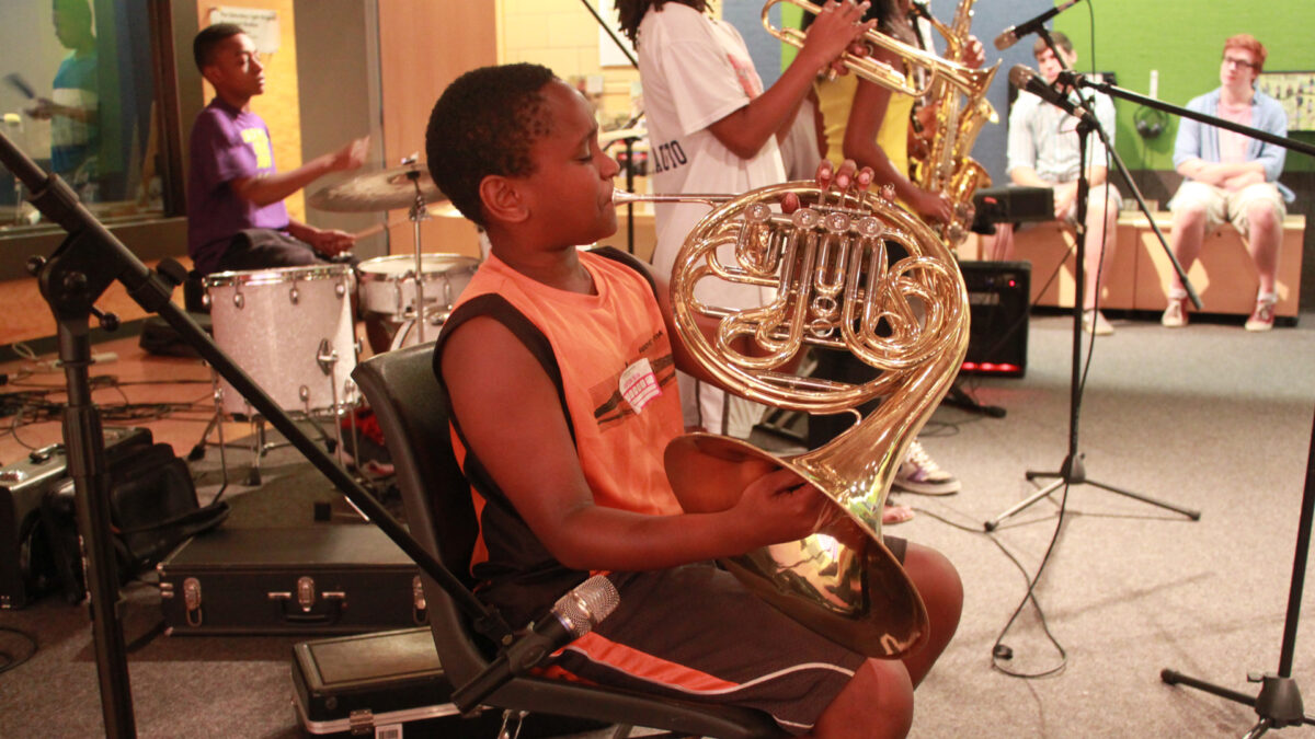 A youth playing a drum set, a youth playing a French horn, a youth playing a trumpet, and a youth playing alto saxophone as two adults watch in the background