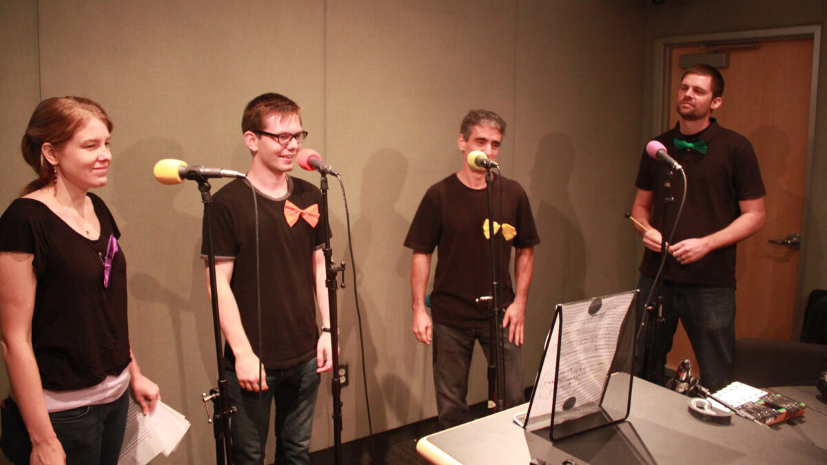 Four adults standing and speaking into microphones in a radio studio