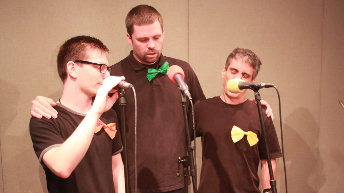 Three adults holding each other's shoulders and speaking into microphones in a radio studio