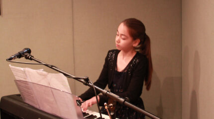 A youth playing a piano keyboard looking at sheet music and sitting behind a microphone in a radio studio