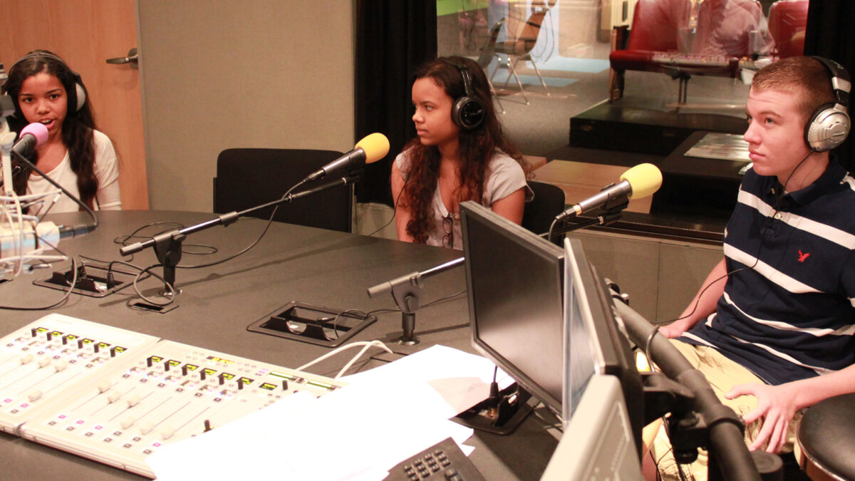 Three teens wearing headphones speaking into microphones in a radio studio