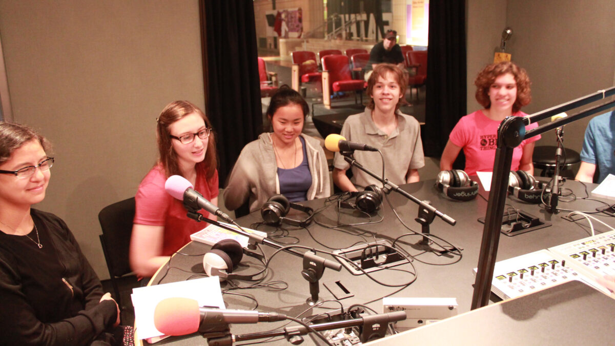 Five teens smiling while sitting behind microphones in a radio studio