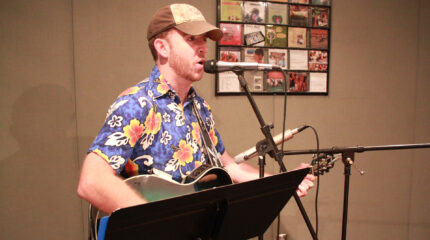 An adult playing an acoustic guitar and singing into a microphone while standing behind a music stand in a radio studio