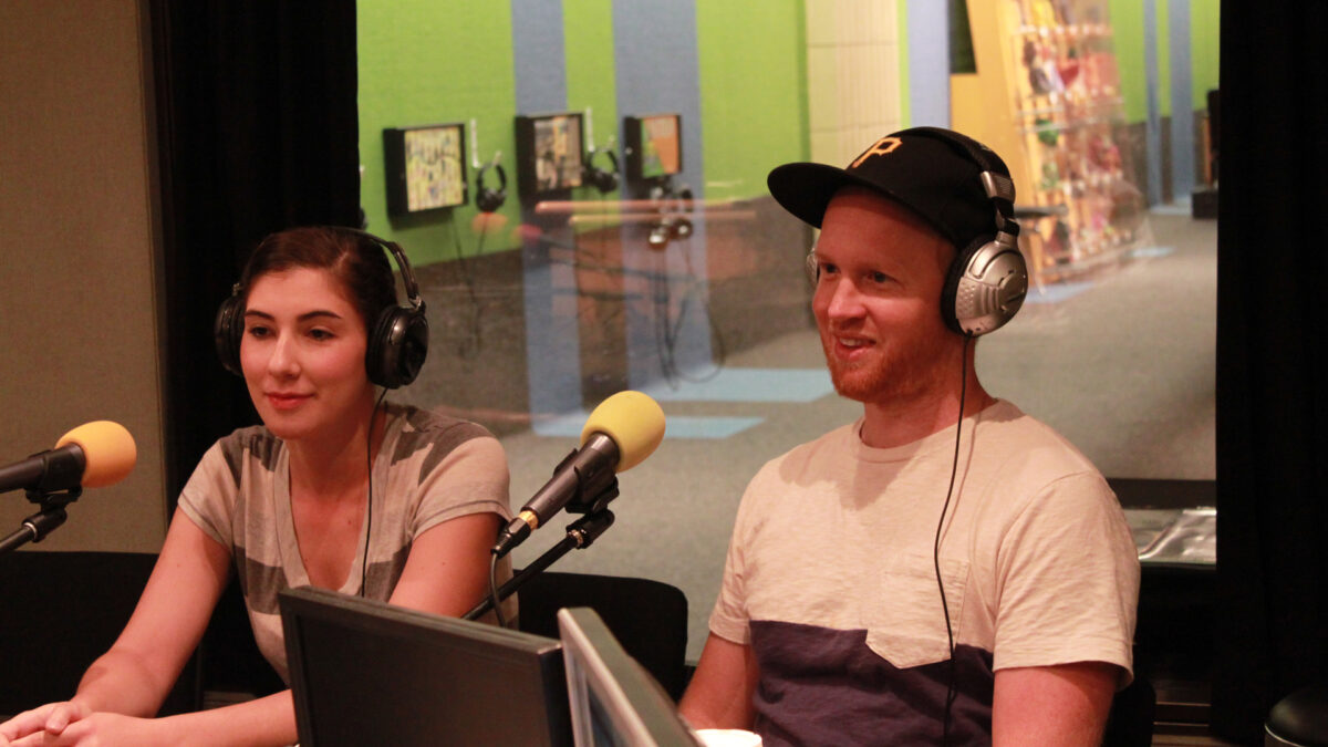 Two adults wearing headphones speaking into microphones in a radio studio